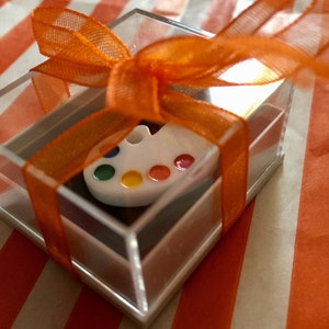 Puede incluir: Una pequeña paleta de artista blanca con puntos de pintura coloridos se presenta en una caja cuadrada transparente. La caja está atada con una cinta naranja brillante, sobre un fondo a rayas naranja y blanco. Regalo para artistas.