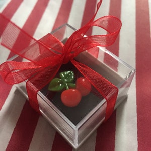 May include: A small clear plastic box with a red ribbon tied around it. Inside the box is a small red cherry with a green stem and leaf.