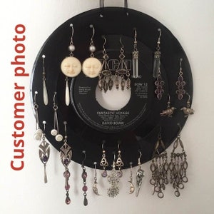 May include: A black vinyl record with holes punched around the edge, used as a jewelry organizer.  Earrings are displayed on the record, including silver, gold, and white earrings with various designs.  The record is labeled "FANTASTIC VOYAGE DAVID BOWIE".
