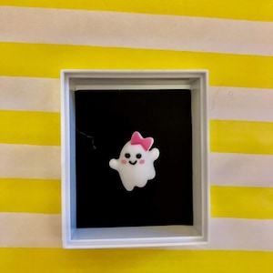 Könnte beinhalten: Eine kleine, weiße Geisterfigur mit einer rosa Schleife und rosigen Wangen wird in einer weißen Box vor einem schwarzen Hintergrund präsentiert. Die Box steht auf einer gelb-weiß gestreiften Oberfläche.