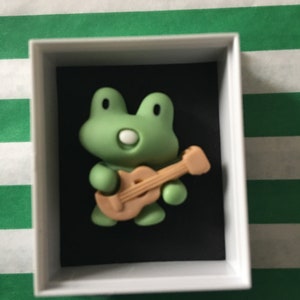 Könnte beinhalten: Eine grüne Froschfigur, die eine braune Gitarre spielt. Die Figur befindet sich in einer weißen Schachtel mit schwarzem Hintergrund.