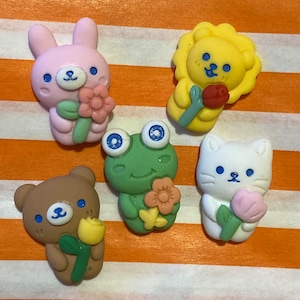 May include: Five colorful ceramic figurines of animals holding flowers. The figurines are a pink bunny, a yellow lion, a green frog, a brown bear, and a white cat. Each figurine is holding a flower with a different color and design.
