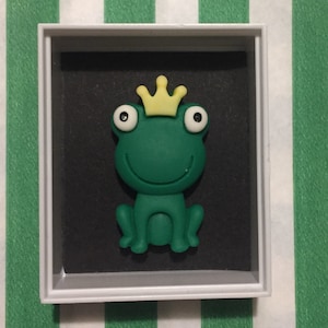 May include: A green frog figurine wearing a yellow crown, sitting in a white frame with a black background.