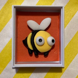 May include: A small, framed picture of a yellow and black bee with white wings and large, white eyes. The bee is on an orange background.