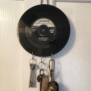 May include: A key holder made from a black vinyl record, featuring the label "LONDON AMERICAN RECORDINGS". The record has three hooks holding keys and keychains. It is suspended by a grey ribbon. The record's label also reads "NO TRESPASSING THE VENTURES".