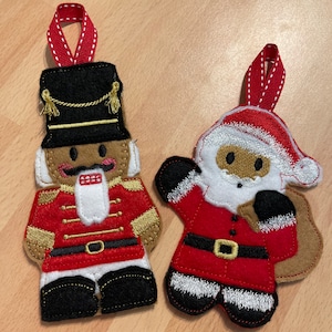 May include: Two felt Christmas decorations, one is a nutcracker in a red and gold uniform and the other is Santa Claus in a red suit with a white beard and a sack.