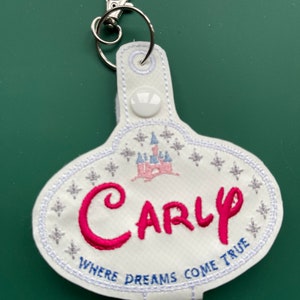 May include: White oval keychain with a pink castle and the name "Carly" embroidered in pink. The text "Where Dreams Come True" is embroidered in blue below the name.