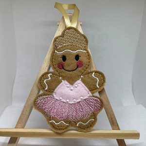 May include: A gingerbread person ornament with a pink tutu and a smiling face. The ornament is made of felt and has a gold ribbon for hanging.