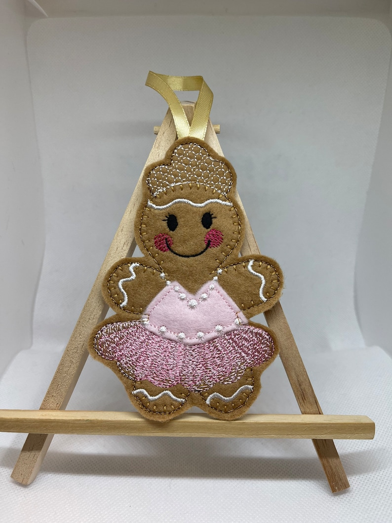 May include: A gingerbread person ornament with a pink tutu and a smiling face. The ornament is made of felt and has a gold ribbon for hanging.