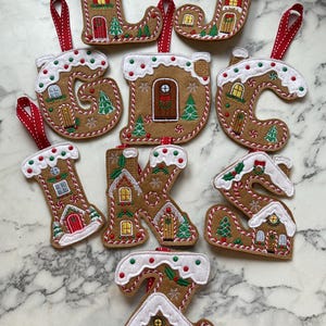 May include: A selection of gingerbread-style Christmas letter ornaments. Each brown felt letter is decorated with white icing, red and green details, and a red ribbon. The letters feature miniature houses and Christmas trees, perfect for festive decor.