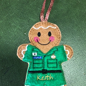 May include: A gingerbread man ornament wearing a green uniform, a name tag that says "Keith", and a brown ribbon for hanging.