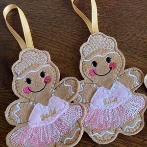 May include: Two gingerbread girl ornaments with pink tutus and gold ribbon hangers. The tutus are embroidered with the names "Bella" and "Ellie".