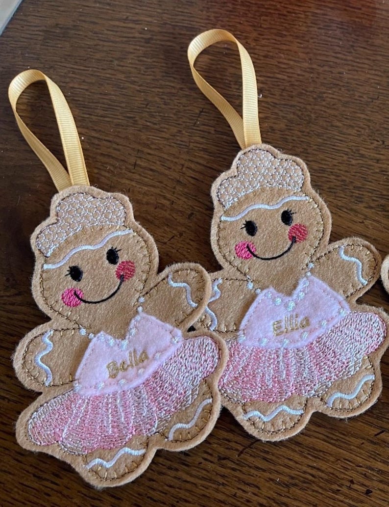 May include: Two gingerbread girl ornaments with pink tutus and gold ribbon hangers. The tutus are embroidered with the names "Bella" and "Ellie".