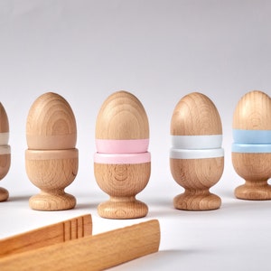 Peut inclure: Six coupelles à œufs en bois avec des bandes peintes en blanc, rose, bleu clair et bleu foncé. Les coupelles à œufs sont disposées en rangée sur une surface blanche.