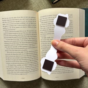 May include: A white paper bookmark with a whimsical design, featuring two black square magnets. The bookmark is held open in a book, with text visible on the pages. The bookmark's shape is irregular, adding to its unique appeal.