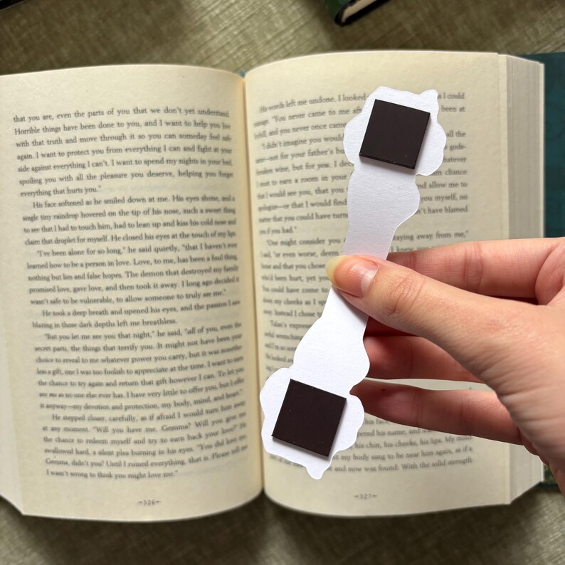 May include: A white paper bookmark with a whimsical design, featuring two black square magnets. The bookmark is held open in a book, with text visible on the pages. The bookmark's shape is irregular, adding to its unique appeal.