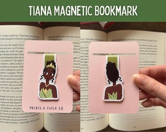Puede incluir: Marcapáginas magnético con una ilustración de Tiana. El marcapáginas tiene un respaldo rosa con una parte superior verde y blanca. La ilustración de Tiana es un dibujo animado de una mujer con un vestido verde y una corona dorada. El texto "PRINTS N PAPER CO" es visible.