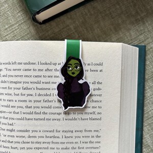 May include: A green and black bookmark featuring a cartoon illustration of a character with green skin, glasses, and a black dress. The bookmark is inserted into a book with white pages and black text. The top of the bookmark is green.