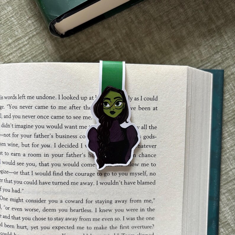 May include: A green and black bookmark featuring a cartoon illustration of a character with green skin, glasses, and a black dress. The bookmark is inserted into a book with white pages and black text. The top of the bookmark is green.