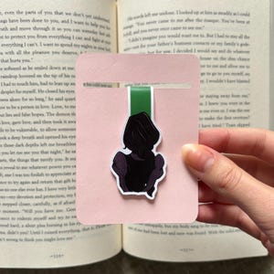 May include: A pink bookmark with a green ribbon and a sticker of a person's silhouette. The person is wearing a dark purple dress. The bookmark is placed in an open book.