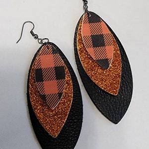 May include: A pair of layered earrings with a plaid pattern. The earrings feature a black, orange glitter, and plaid design. The earrings have a fish hook style closure and are designed to be worn on the ear.