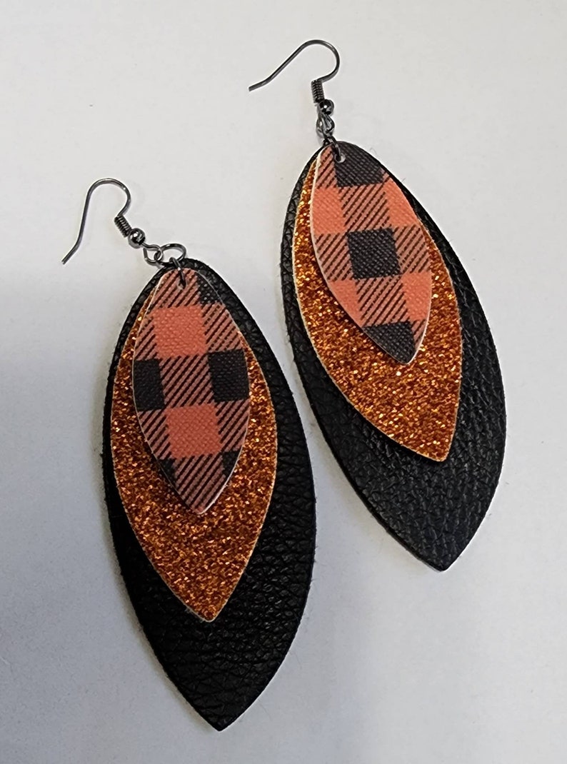 May include: A pair of layered earrings with a plaid pattern. The earrings feature a black, orange glitter, and plaid design. The earrings have a fish hook style closure and are designed to be worn on the ear.