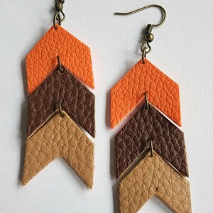 May include: A pair of layered leather earrings in an arrow design. The earrings feature three layers of textured leather in orange, brown, and tan colors. The earrings have bronze-colored ear wires.