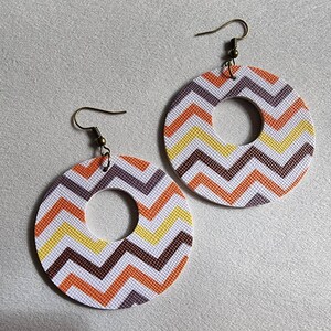 May include: A pair of hoop earrings with a chevron pattern in white, orange, yellow, and brown. The earrings have a circular design with a cutout in the center and bronze-colored ear wires.