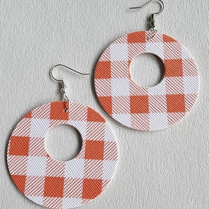 May include: A pair of orange and white plaid hoop earrings. The earrings are circular with a smaller circle cut out of the center. The earrings hang from silver-colored fish hook ear wires. The plaid pattern is a classic gingham design.