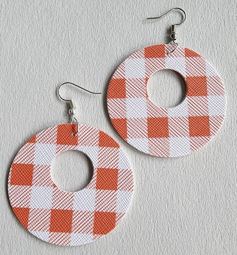 May include: A pair of orange and white plaid hoop earrings. The earrings are circular with a smaller circle cut out of the center. The earrings hang from silver-colored fish hook ear wires. The plaid pattern is a classic gingham design.