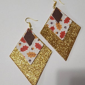 May include: A pair of diamond-shaped earrings. The earrings feature a gold glitter base, a layer with a fall leaf pattern, and a small brown diamond at the top. The earrings have gold-colored fish hook ear wires.