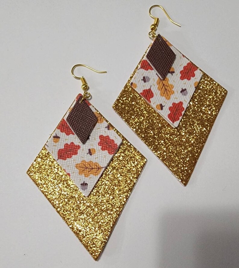 May include: A pair of diamond-shaped earrings. The earrings feature a gold glitter base, a layer with a fall leaf pattern, and a small brown diamond at the top. The earrings have gold-colored fish hook ear wires.