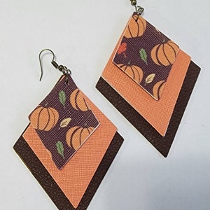 May include: A pair of layered diamond-shaped earrings. The top layer features a pumpkin pattern on a dark purple background. The middle layer is orange, and the bottom layer is dark brown. The earrings have bronze-colored hooks.