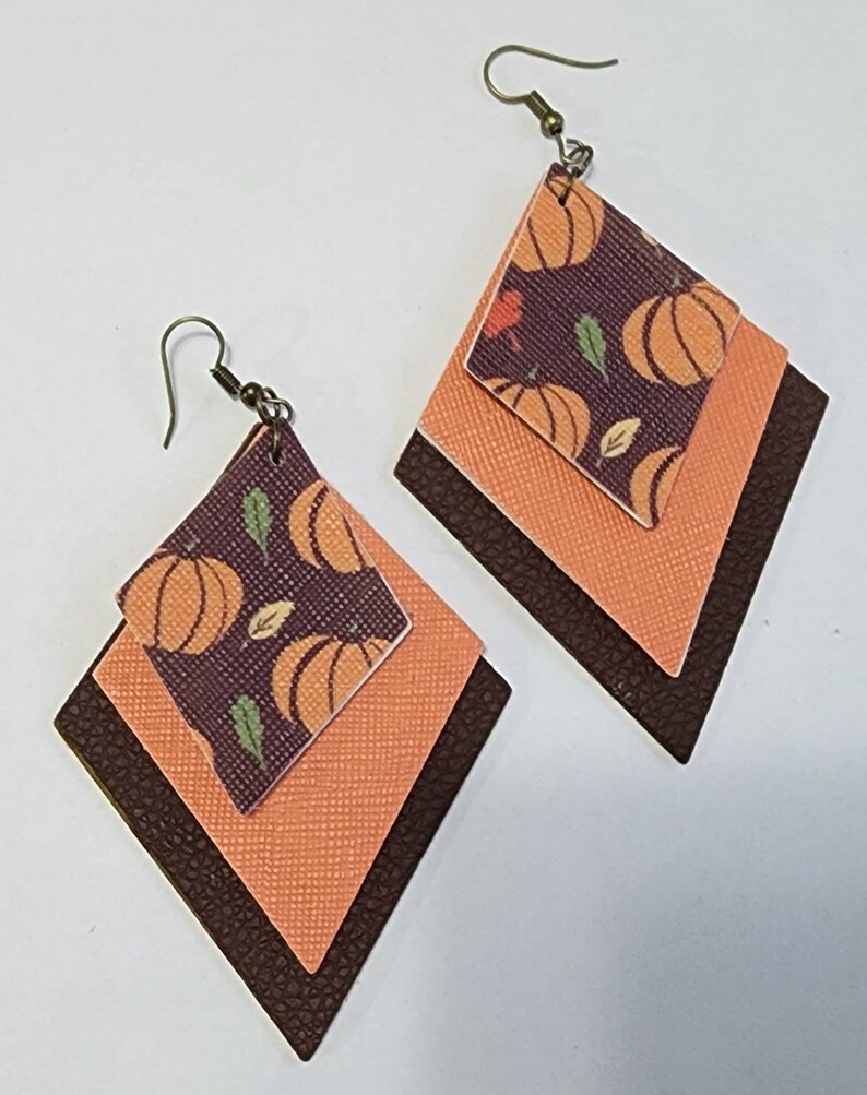 May include: A pair of layered diamond-shaped earrings. The top layer features a pumpkin pattern on a dark purple background. The middle layer is orange, and the bottom layer is dark brown. The earrings have bronze-colored hooks.