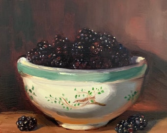 Puede incluir: Un tazón de cerámica blanca con un borde verde y un diseño floral, lleno de moras maduras. El tazón está sentado sobre una superficie de madera con dos moras adicionales a su lado.