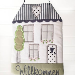 May include: A white house-shaped door hanger with a green tree, grey windows, and a black and white polka dot door. The door has a small black heart on it. The top of the house is grey and white striped with a grey heart with white polka dots. The bottom of the house has a green border with the word "Willkommen" in black.