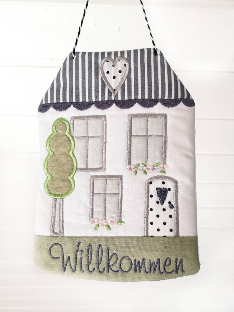 May include: A white house-shaped door hanger with a green tree, grey windows, and a black and white polka dot door. The door has a small black heart on it. The top of the house is grey and white striped with a grey heart with white polka dots. The bottom of the house has a green border with the word "Willkommen" in black.