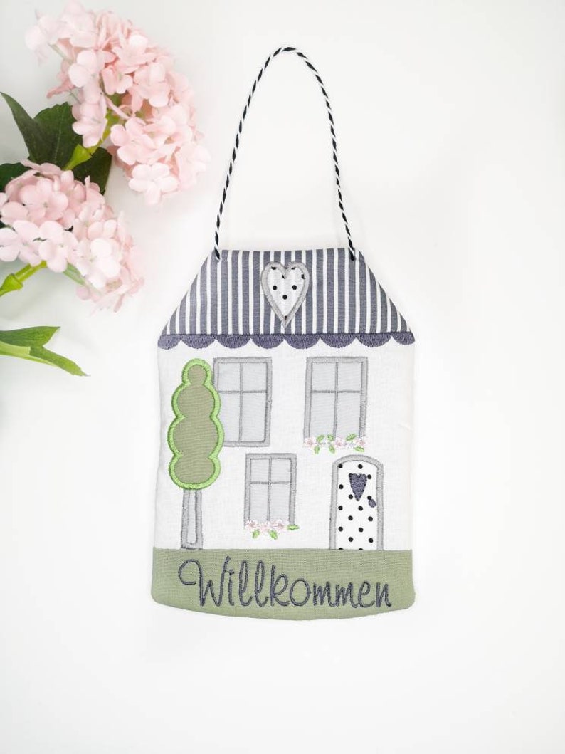 May include: A white house-shaped door hanger with a grey and white striped roof, a green tree, and a black and white polka dot door. The door has a small heart-shaped window. The hanger has a grey embroidered welcome sign that says "Willkommen".