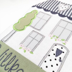 May include: A white house-shaped doorstop with a green tree, grey windows, and a black and white polka dot door. The doorstop has a green fabric banner with the word "welcome" embroidered in grey.