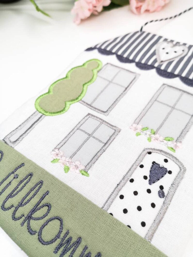 May include: A white house-shaped doorstop with a green tree, grey windows, and a black and white polka dot door. The doorstop has a green fabric banner with the word "welcome" embroidered in grey.