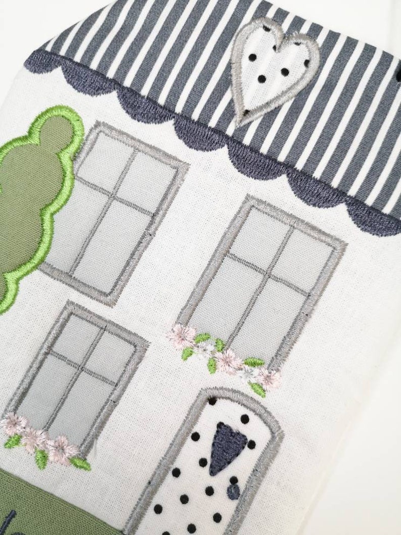 May include: A white embroidered house with a grey and white striped roof, a green bush, a grey door with black polka dots, and two windows with grey frames.