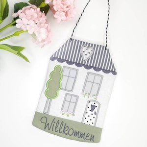 May include: A white house-shaped door hanger with a green tree, grey windows, and a black and white polka dot door. The door has a small heart on it. The bottom of the hanger has a green border with the word "Willkommen" in grey embroidery.