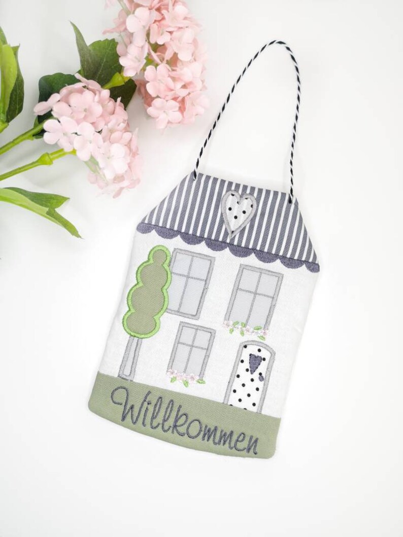 May include: A white house-shaped door hanger with a green tree, grey windows, and a black and white polka dot door. The door has a small heart on it. The bottom of the hanger has a green border with the word "Willkommen" in grey embroidery.