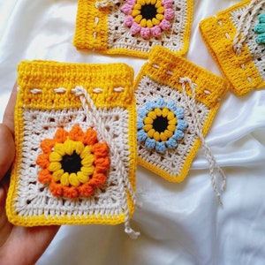 Op de afbeelding: Vijf gehaakte tasjes met zonnebloemmotieven in verschillende kleuren, waaronder geel, roze, blauw en paars. Elk tasje heeft een trekkoord en is gemaakt van wit en geel garen.