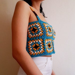Pode incluir: Um top de crochê azul, branco e marrom com um padrão quadrado. O top é sem mangas e tem um comprimento curto.