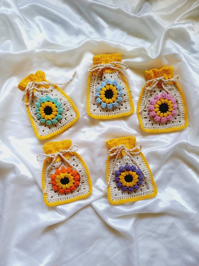 Op de afbeelding: Vijf gehaakte zonnebloemvormige zakjes met gele trekkoordjes. De zakjes zijn in verschillende kleuren, waaronder roze, blauw, paars en oranje.