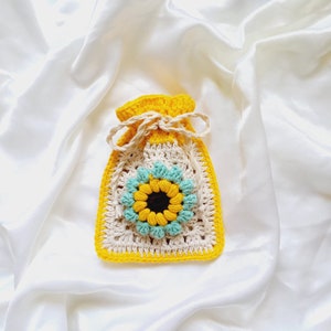 Op de afbeelding: Een gehaakte tas in de vorm van een zonnebloem met een gele rand, een cr&egrave;mekleurige binnenkant en blauwe bloemblaadjes. De tas ligt op een witte satijnen achtergrond.
