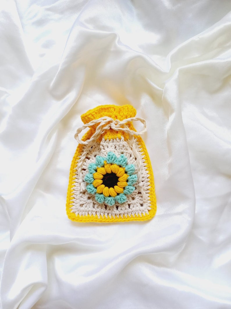 Op de afbeelding: Een gehaakte tas in de vorm van een zonnebloem met een gele rand, een cr&egrave;mekleurige binnenkant en blauwe bloemblaadjes. De tas ligt op een witte satijnen achtergrond.