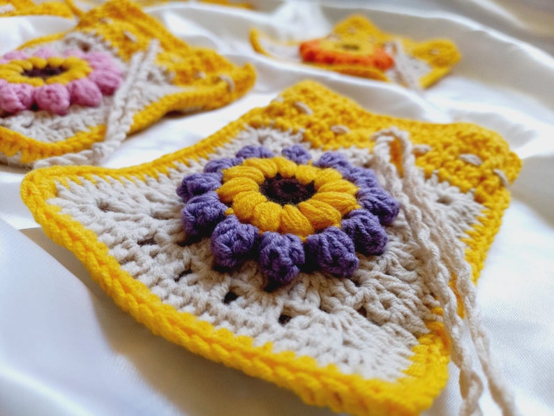 Op de afbeelding: Een gehaakte tas met een gele rand en een bloemmotief in het midden. De bloem heeft een gele kern, paarse bloemblaadjes en een bruine kern.