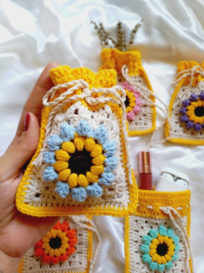 Op de afbeelding: Vijf gehaakte trekkoordzakjes met een zonnebloemdessin in geel, blauw, oranje en wit. De zakjes zijn gemaakt met een granny square-patroon en hebben een beige achtergrond.